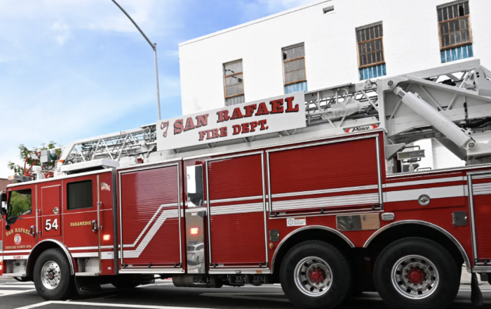 A red San Rafael Fire Department ladder truck with "City of San Rafael Fire Dept." painted on the side, parked on a street in front of a building