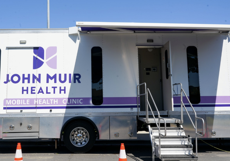 A John Muir Health Mobile Health Clinic truck with its door and steps extended, parked with traffic cones nearby.