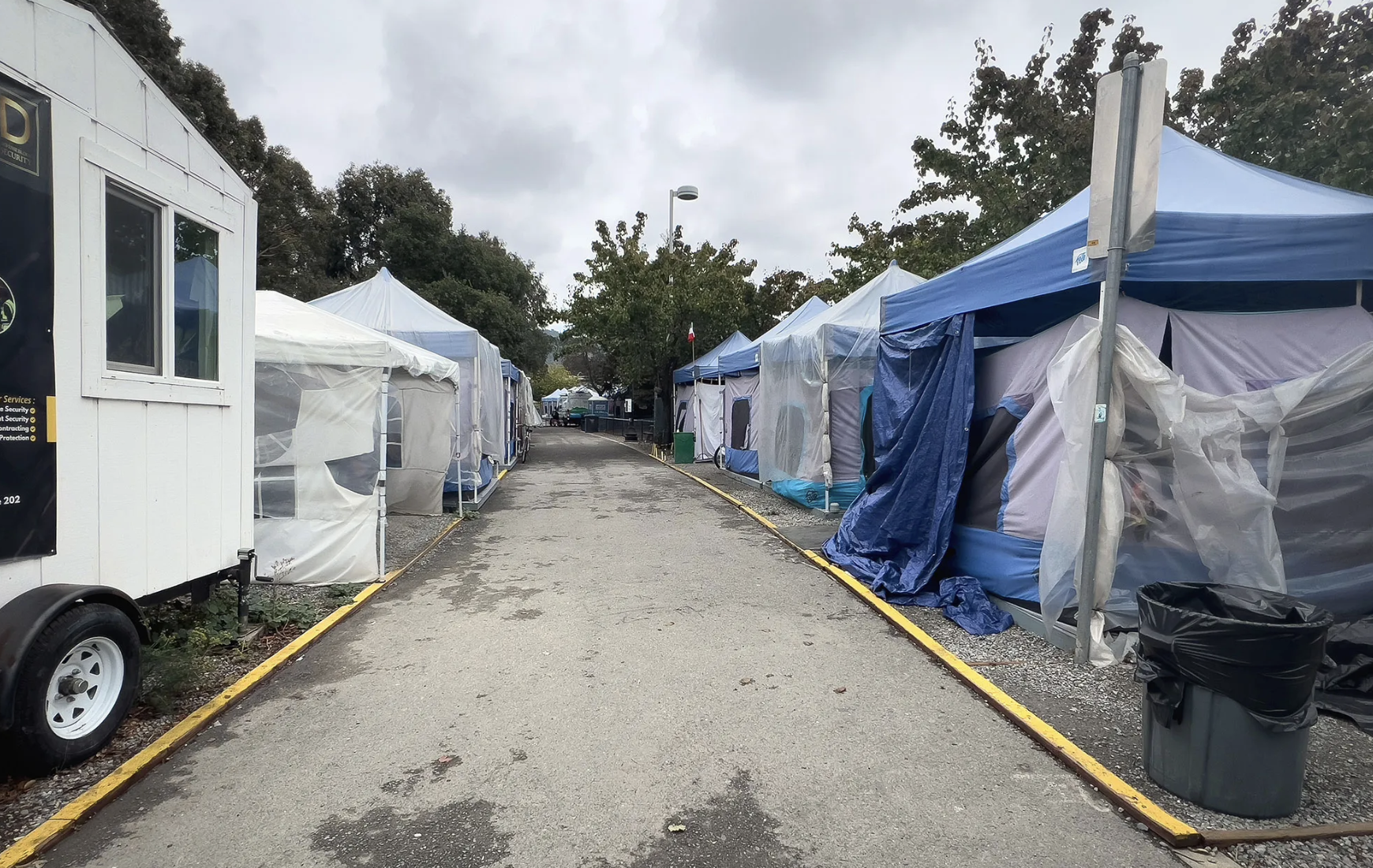 A row of tents and canopy shelters lining a paved path at an outdoor homeless encampment, with a security trailer and trash bins visible.