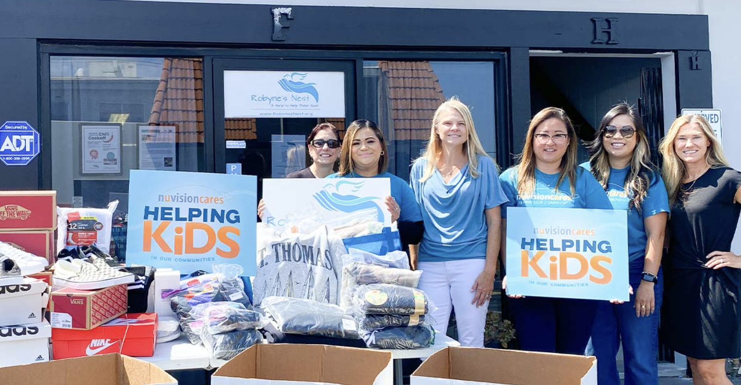 Volunteers from NuVision Credit Union pose with donated shoes, clothing, and supplies outside Robyne's Nest, holding "NuVision Cares: Helping Kids in Our Communities" signs.