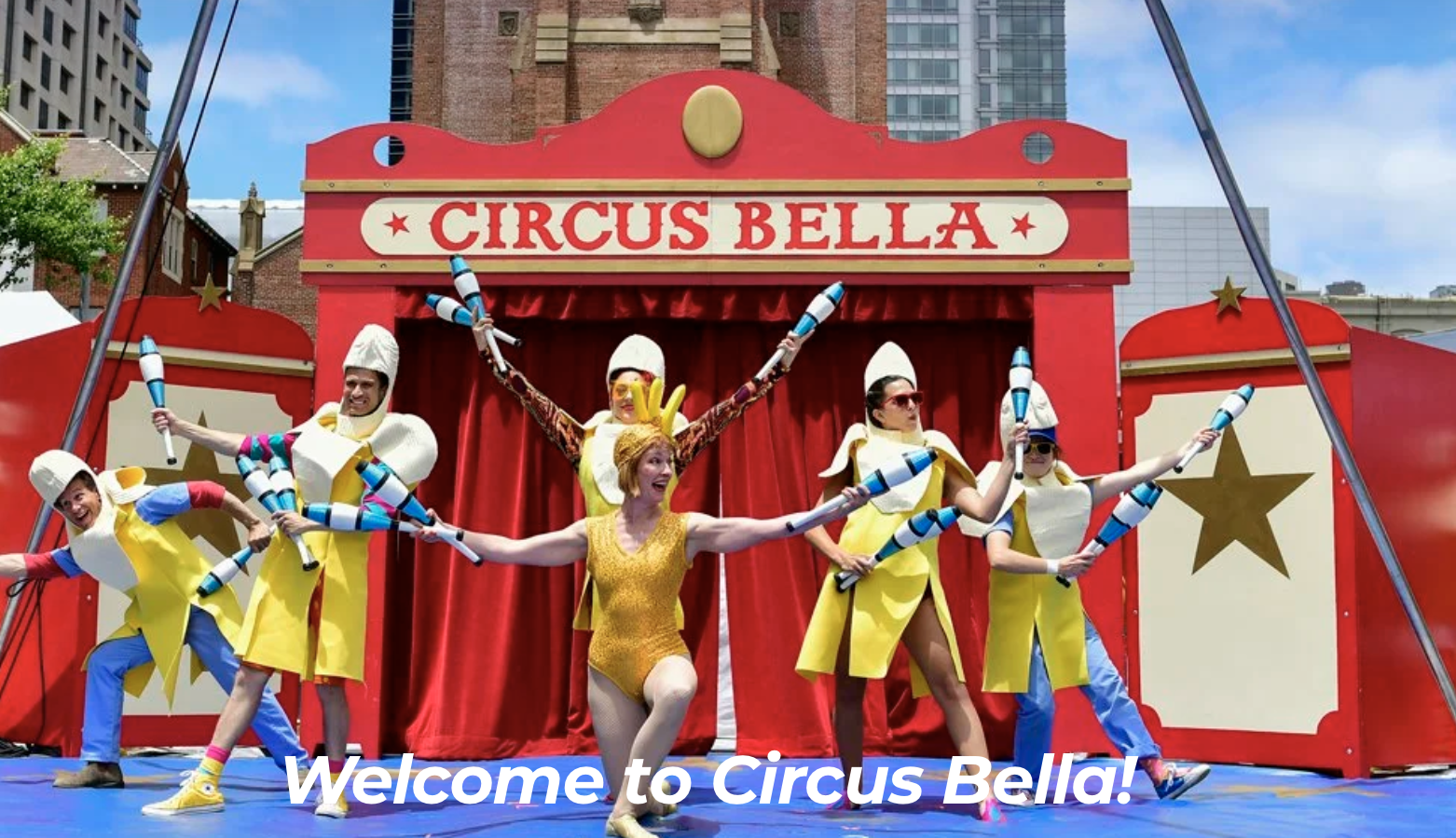 Circus Bella performers in yellow banana costumes hold juggling clubs and pose in front of the troupe's red and gold "Circus Bella" stage backdrop, with a performer in a gold leotard kneeling center stage, text reading "Welcome to Circus Bella!"