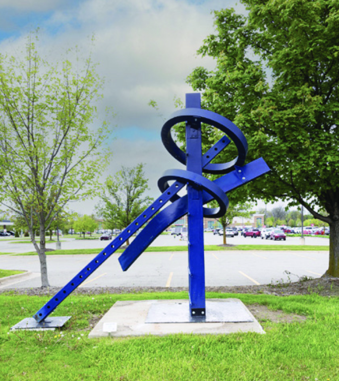 Ray Katz's abstract blue steel sculpture "Ring Walker," featuring circular ring shapes and angled bars, stands on a concrete base near a parking lot.