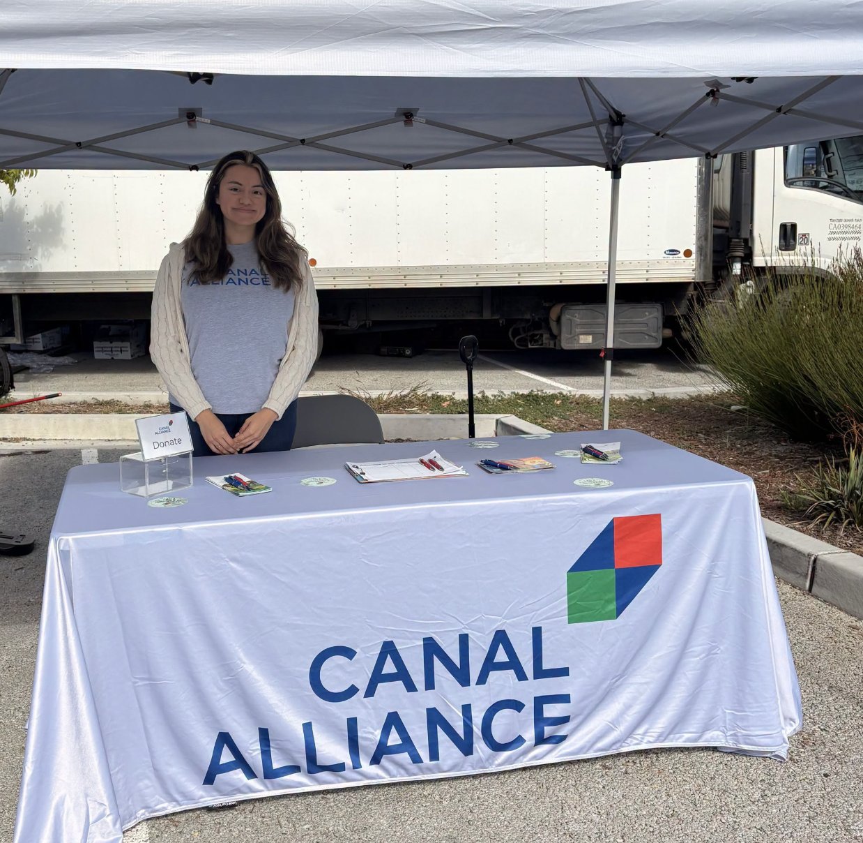 A Canal Alliance volunteer stands behind an outdoor table with the organization's branded tablecloth, donation box, and informational materials at a community event.