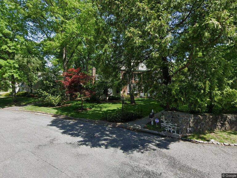 A tree-lined residential street with a brick home, landscaped yard and low stone wall.
