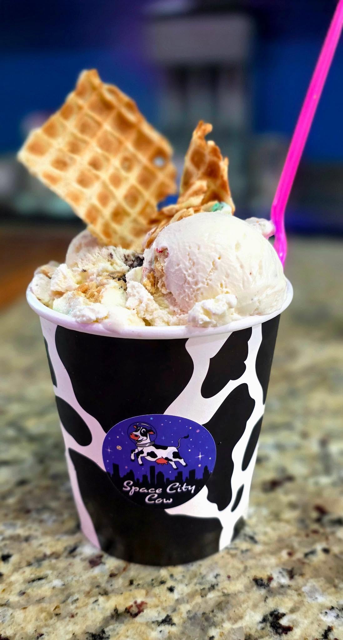 A cup of assorted ice cream topped with waffle pieces and a pink spoon, served in a black-and-white cow-print cup.