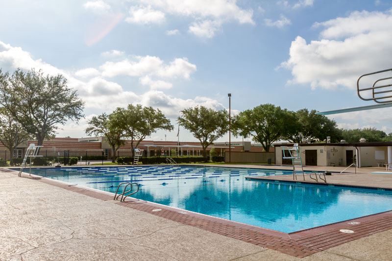 Outdoor swimming pool with clear blue water, lane markings, diving platforms, and a surrounding concrete deck under a partly cloudy sky.