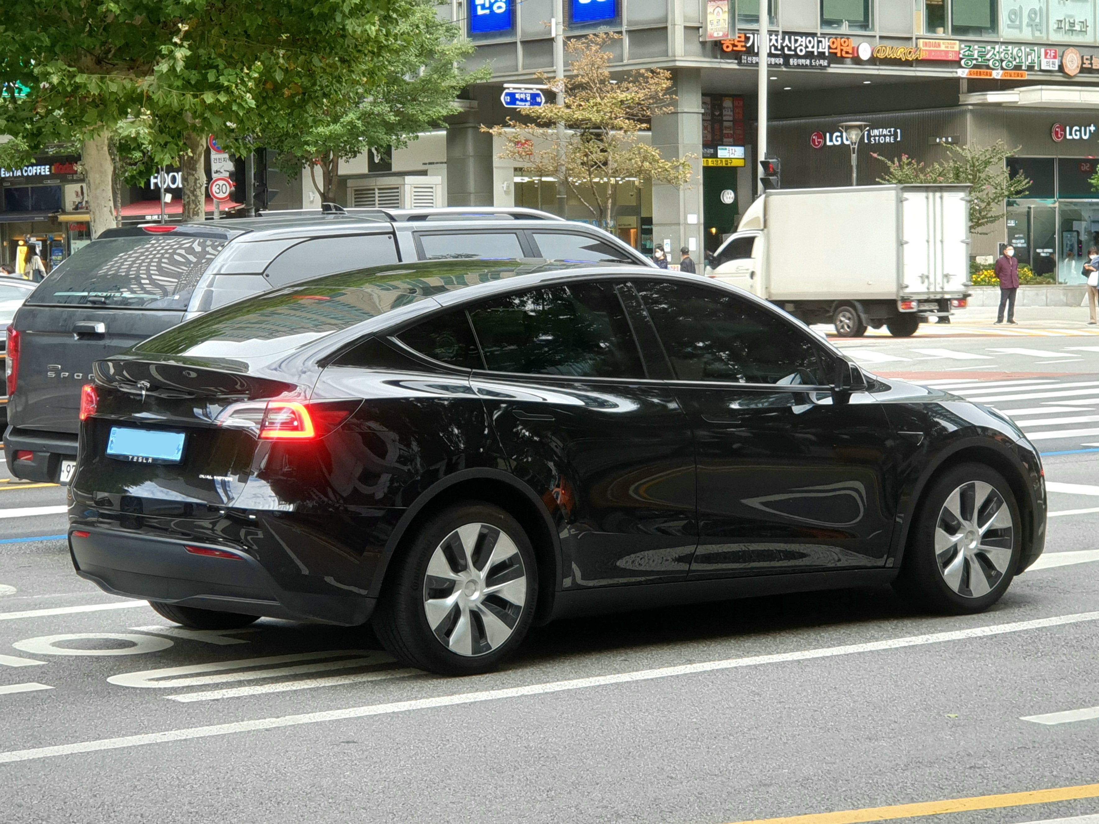 Black Tesla Model Y viewed from the rear and side on a city street, with other vehicles, storefronts and pedestrians in the background.