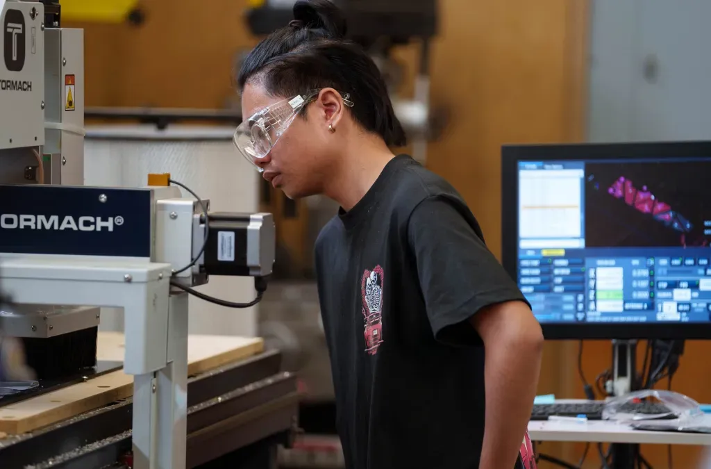A teenage student wearing safety glasses operates a computer-controlled router machine in a high school robotics workshop.