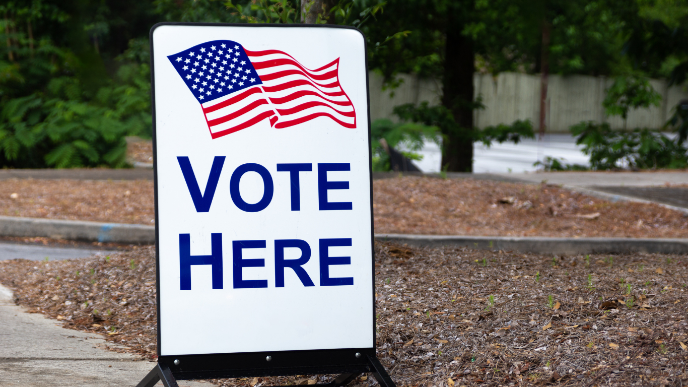 A white “VOTE HERE” sign featuring an American flag graphic, standing beside a sidewalk in a landscaped outdoor area.