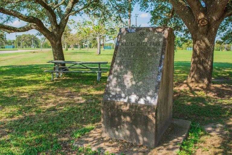 A stone memorial marker stands beneath large trees in a grassy park, with picnic tables and sports fields visible in the background.