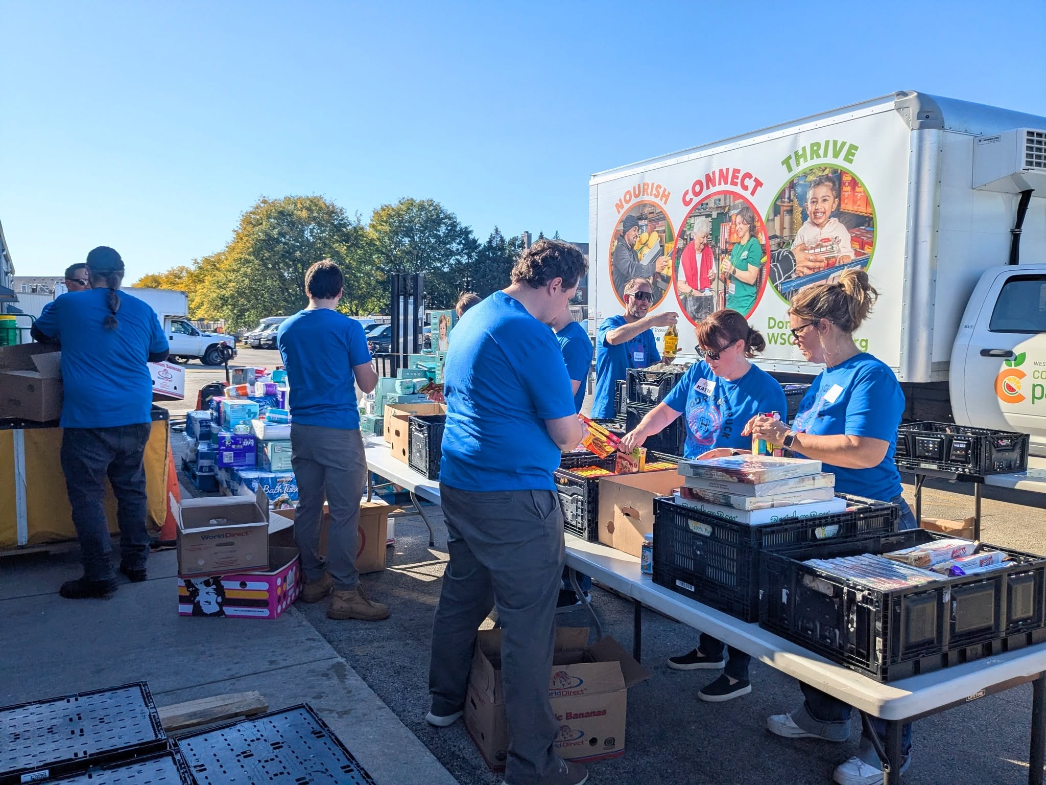 Volunteers in blue shirts sort donated food and household supplies on outdoor tables beside a food pantry truck.