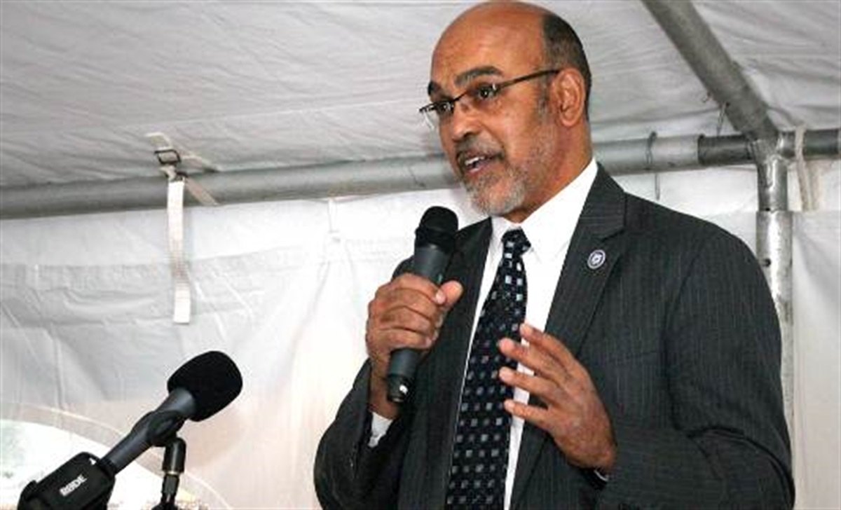 A man in a dark suit and glasses speaks into a microphone at an outdoor podium.