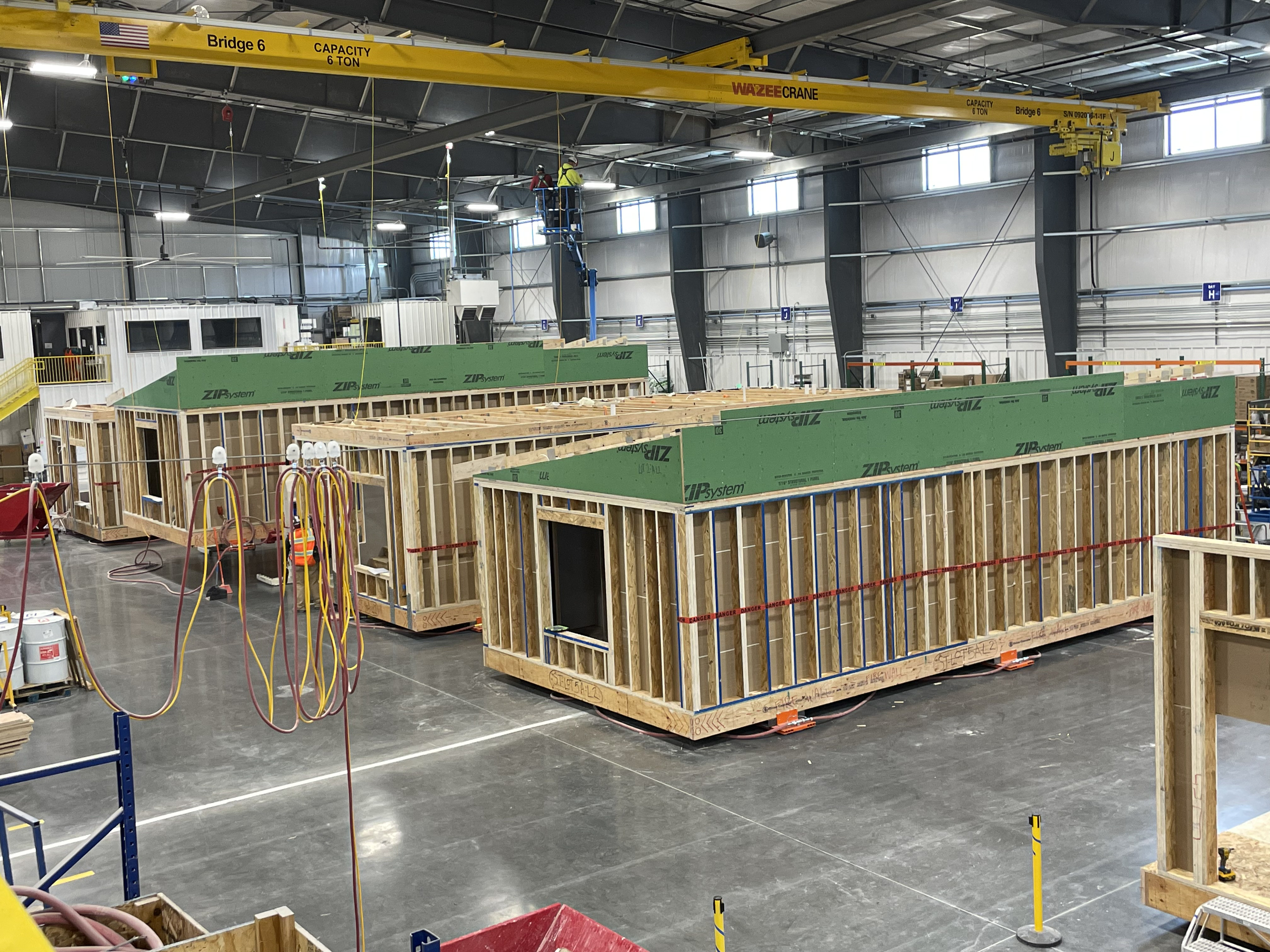 Partially built modular home units on a factory floor with an overhead crane system.