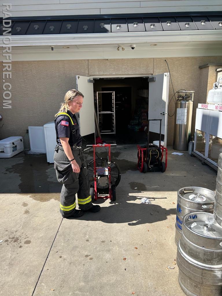 Firefighter in turnout gear standing next to a red wheeled ventilation fan outside a building.