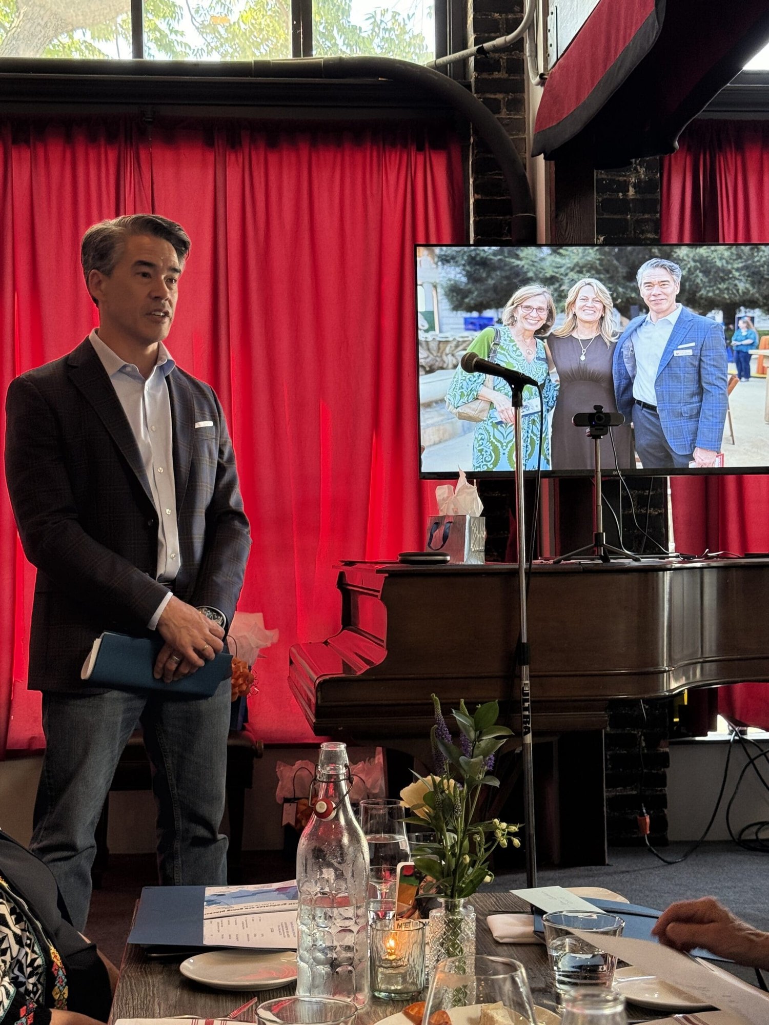 A man in a dark blazer stands beside a TV screen during a presentation at a venue with red curtains and a piano.
