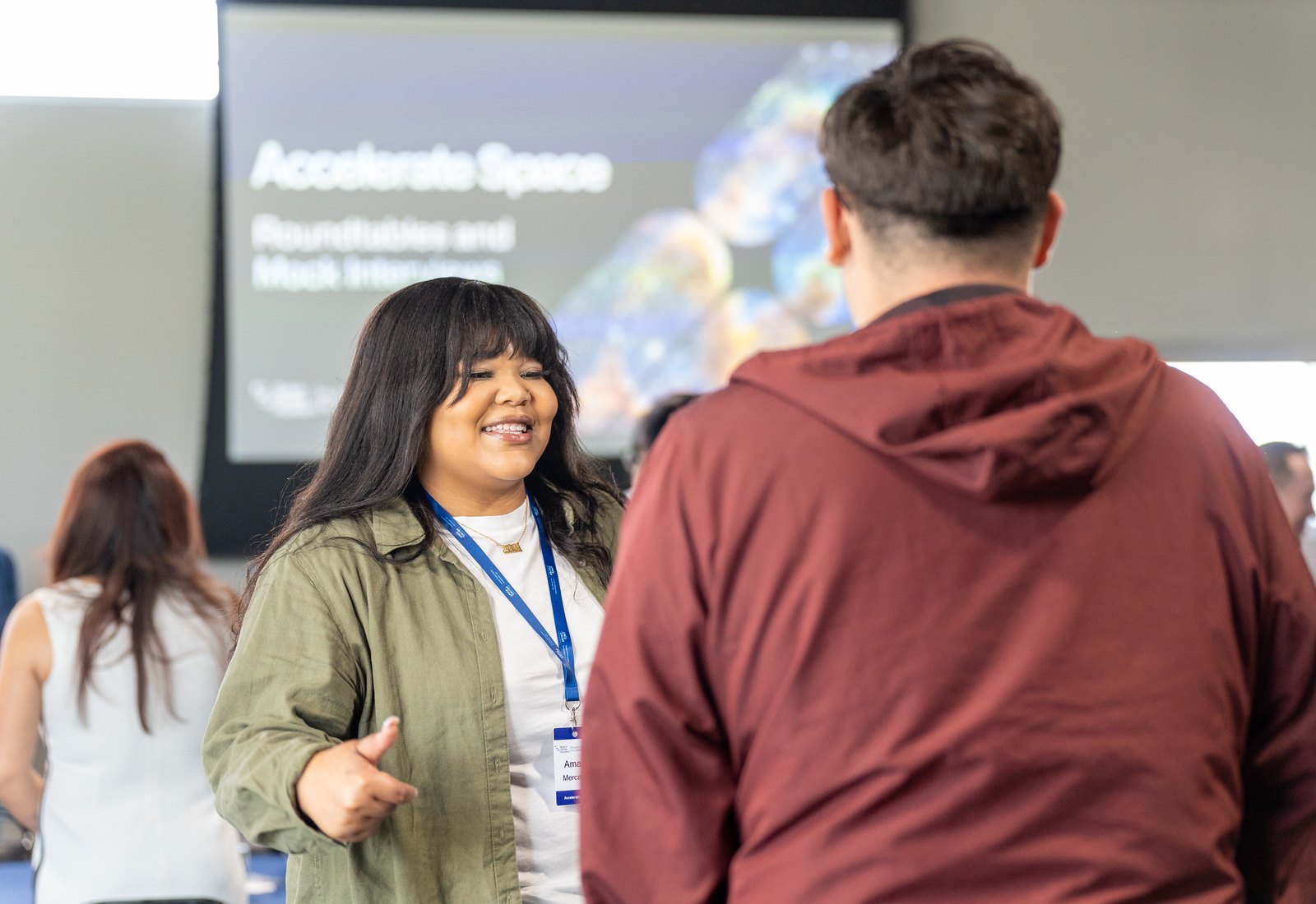 Two people converse at an indoor event with an 'Accelerate Space' presentation slide visible behind them.