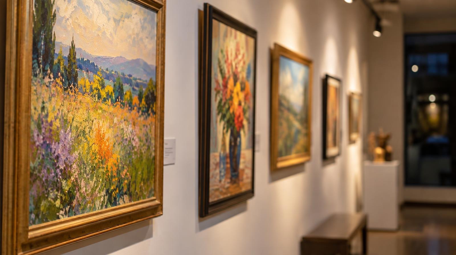 Colorful framed paintings hang on a white gallery wall under warm spotlights.