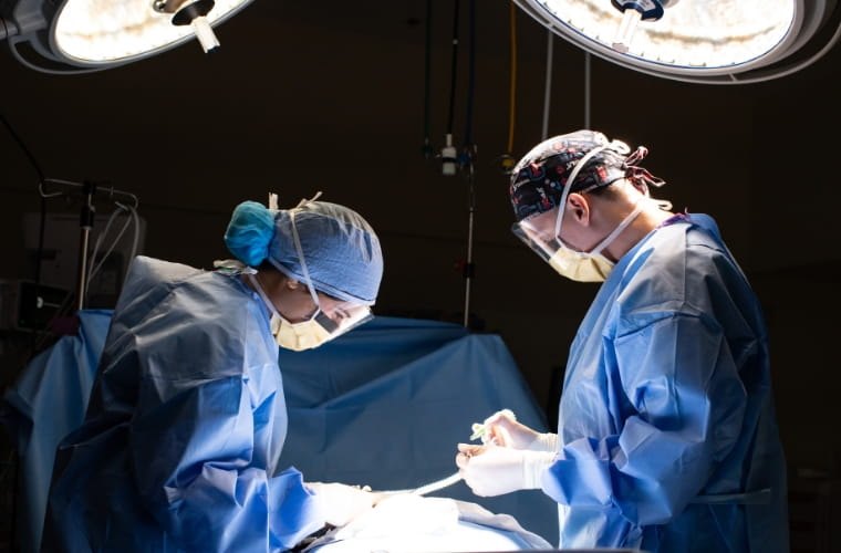 Two surgeons in blue gowns and masks operating under bright overhead lights.