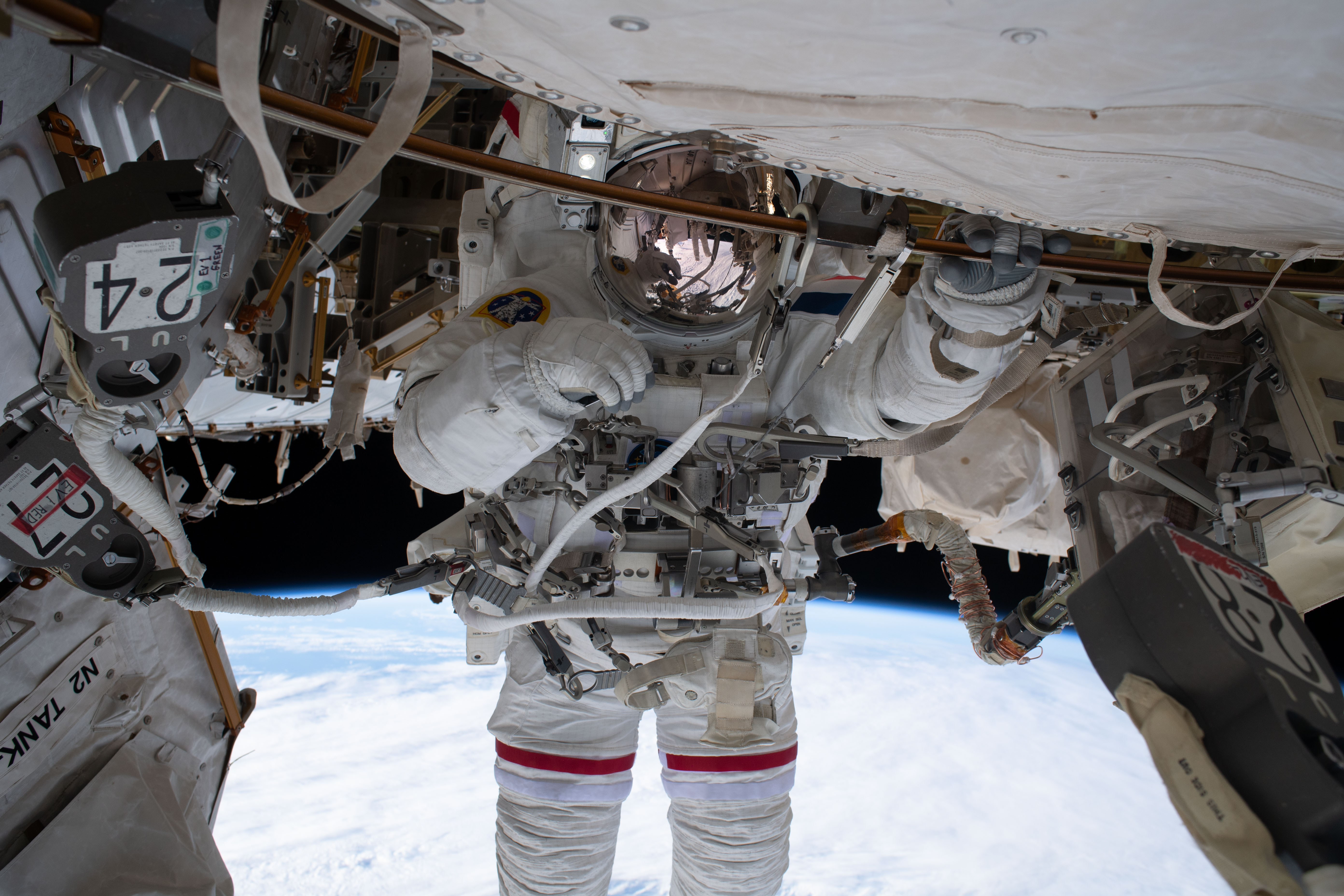 Astronaut in white spacesuit with gold visor tethered to space station exterior, Earth below.