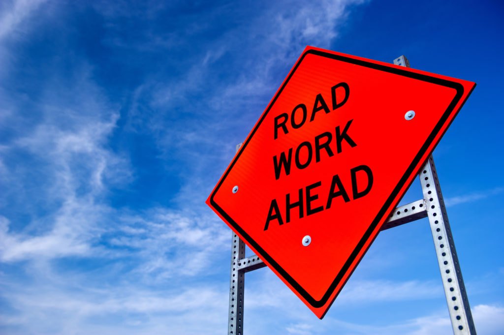 Orange diamond-shaped Road Work Ahead sign on metal posts against a blue sky.