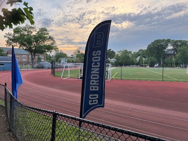 Navy blue Go Broncos feather banner at a track and turf field complex at dusk.