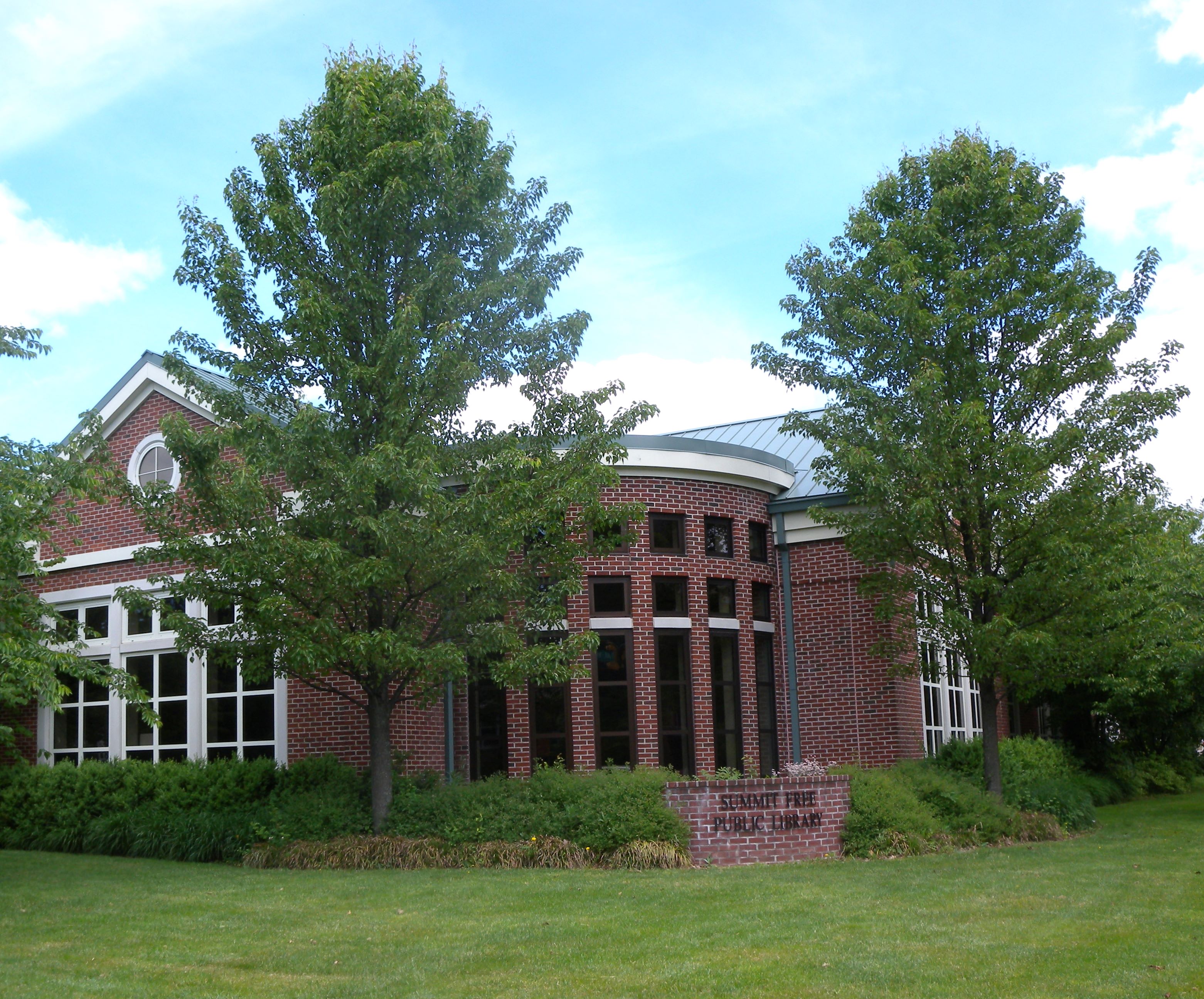 Brick public library building with a curved entrance tower and metal roof, flanked by large trees.