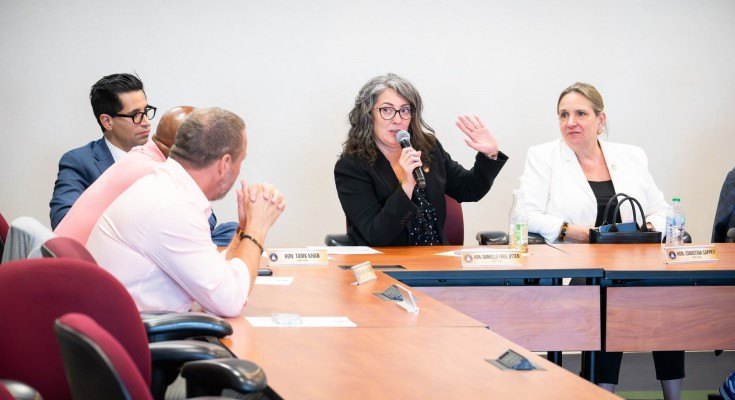 A woman speaks into a microphone at a conference table during a formal roundtable meeting.