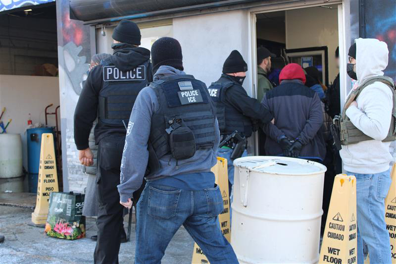 Multiple officers in POLICE ICE vests and tactical gear enter a building.