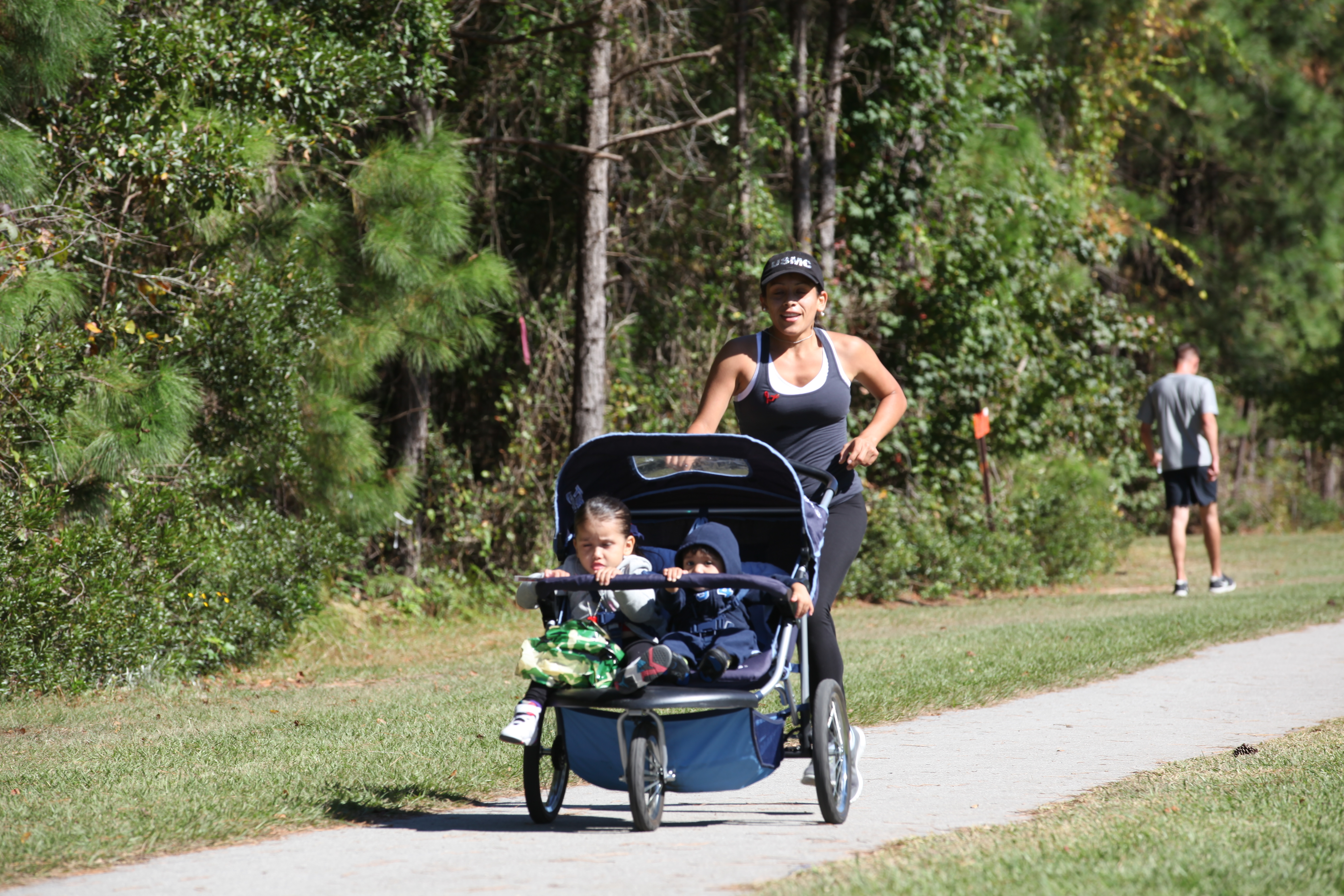 A woman pushes a jogging stroller with two children along a paved, tree-lined trail.