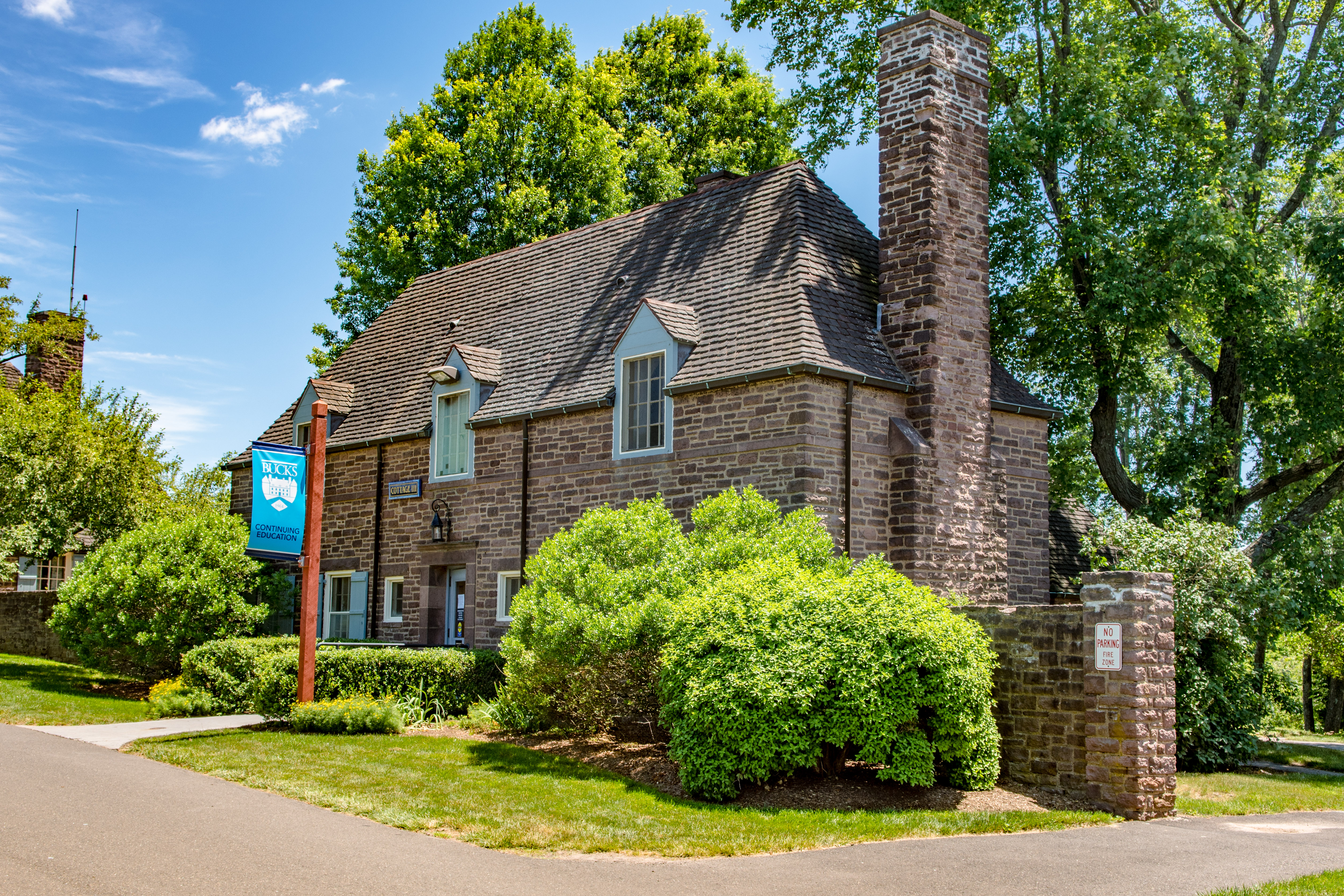 Stone cottage building with steep roof and blue college banner on a sunny campus.