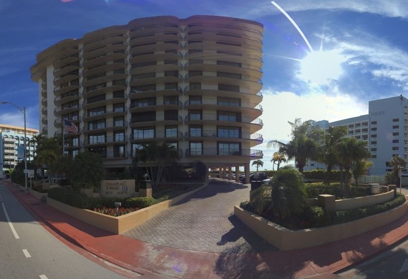 A curved 12-story beachfront condominium building with palm trees and a driveway entrance under a blue sky.