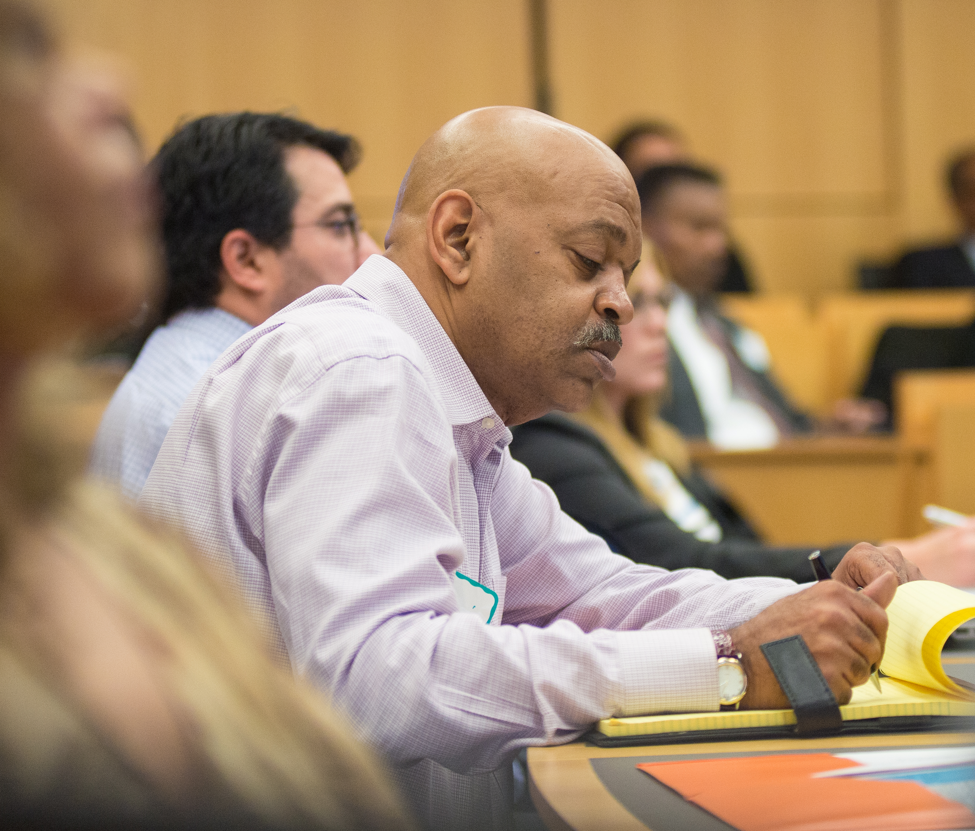 A man in a checkered shirt leans over a yellow notepad taking notes at a workshop.