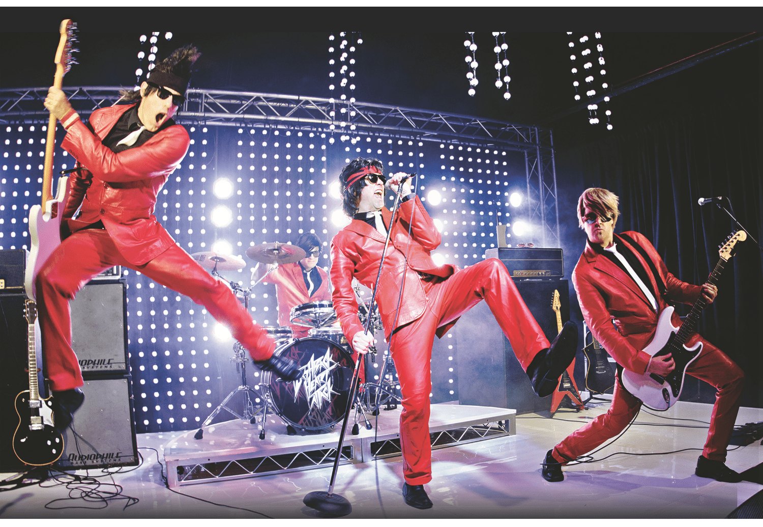Four performers in matching red suits playing guitars and singing on a lit stage.