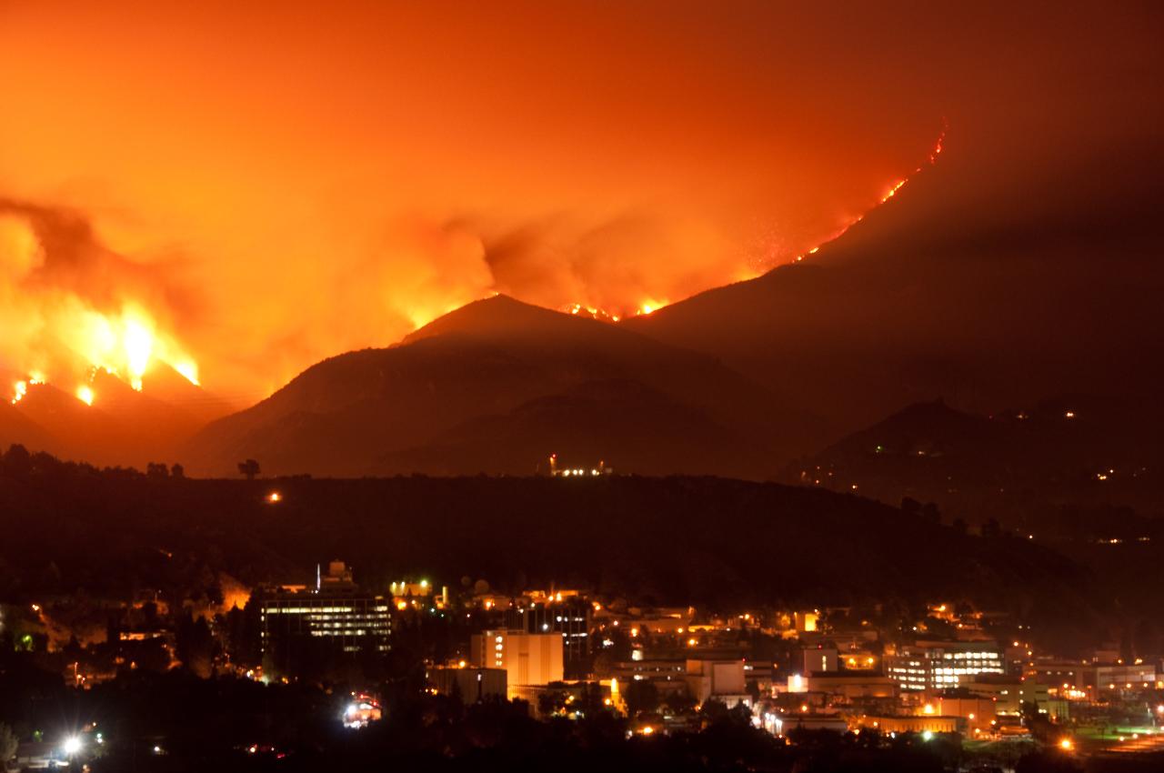Wildfire burns across dark hillsides at night, sky glowing orange-red above city lights below.