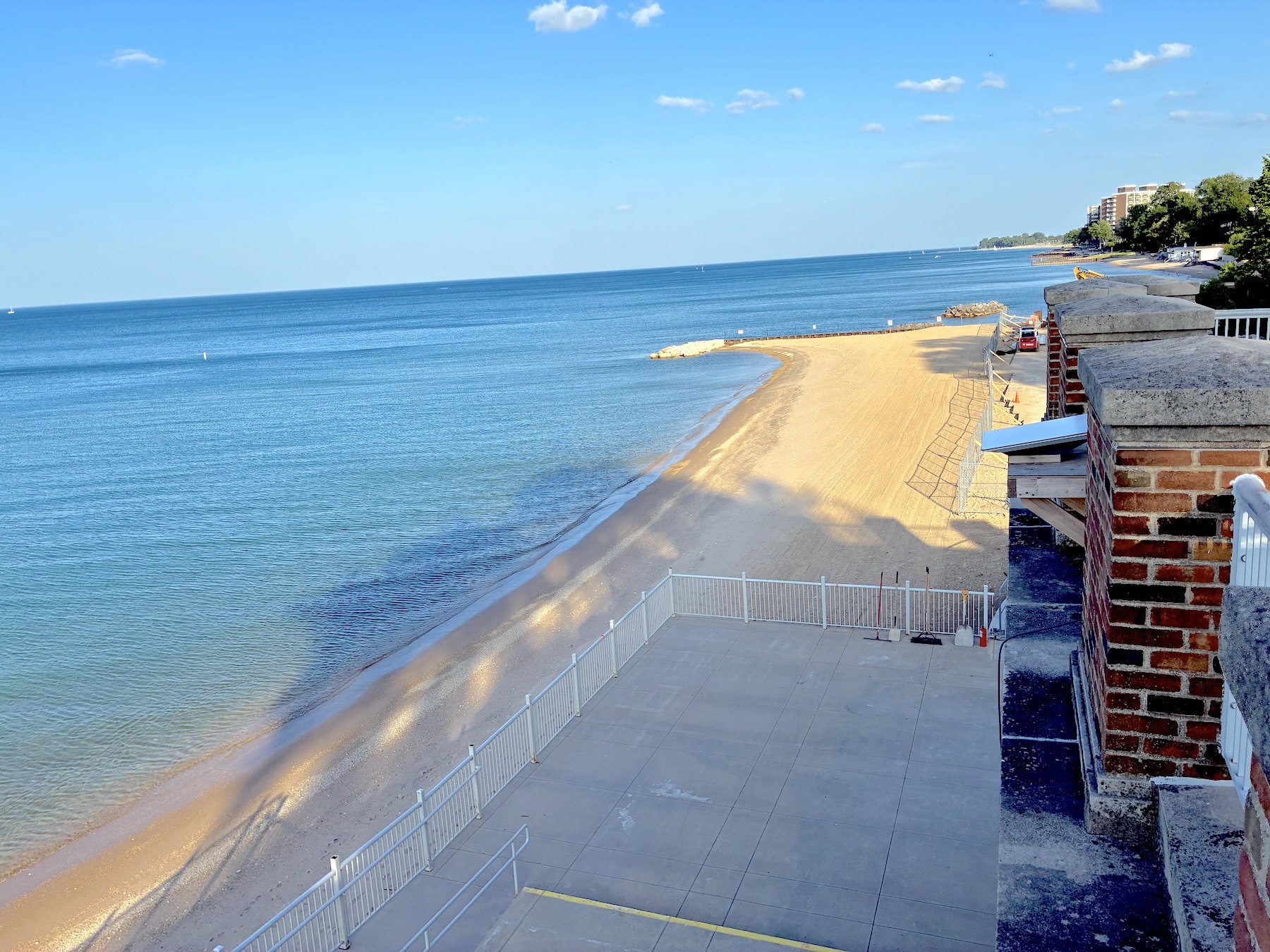 Sandy beach along calm lake waters with a concrete plaza, white metal fencing, and brick structures under a clear blue sky.