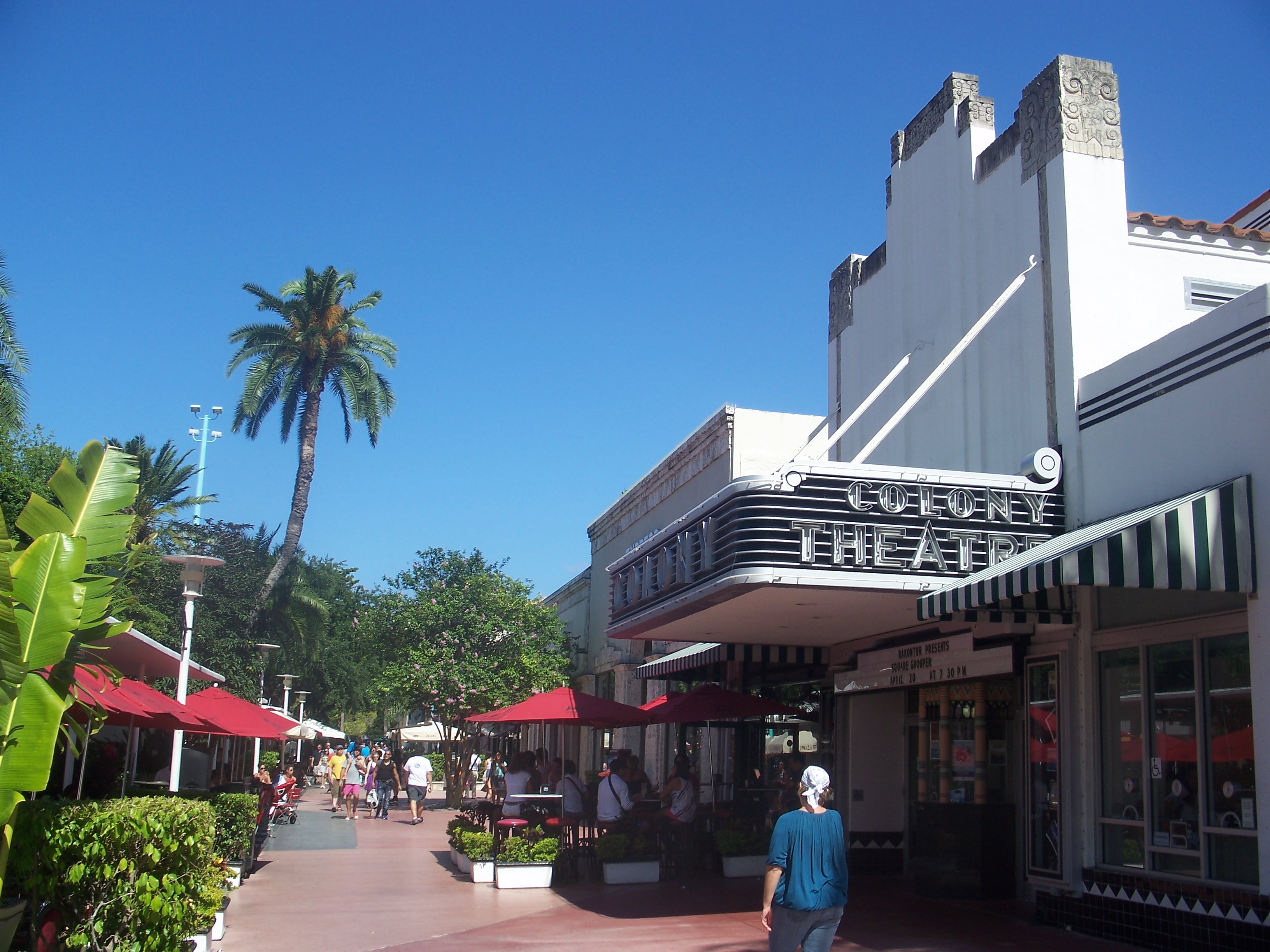 Art Deco theater facade with neon marquee sign on a palm-lined pedestrian mall.