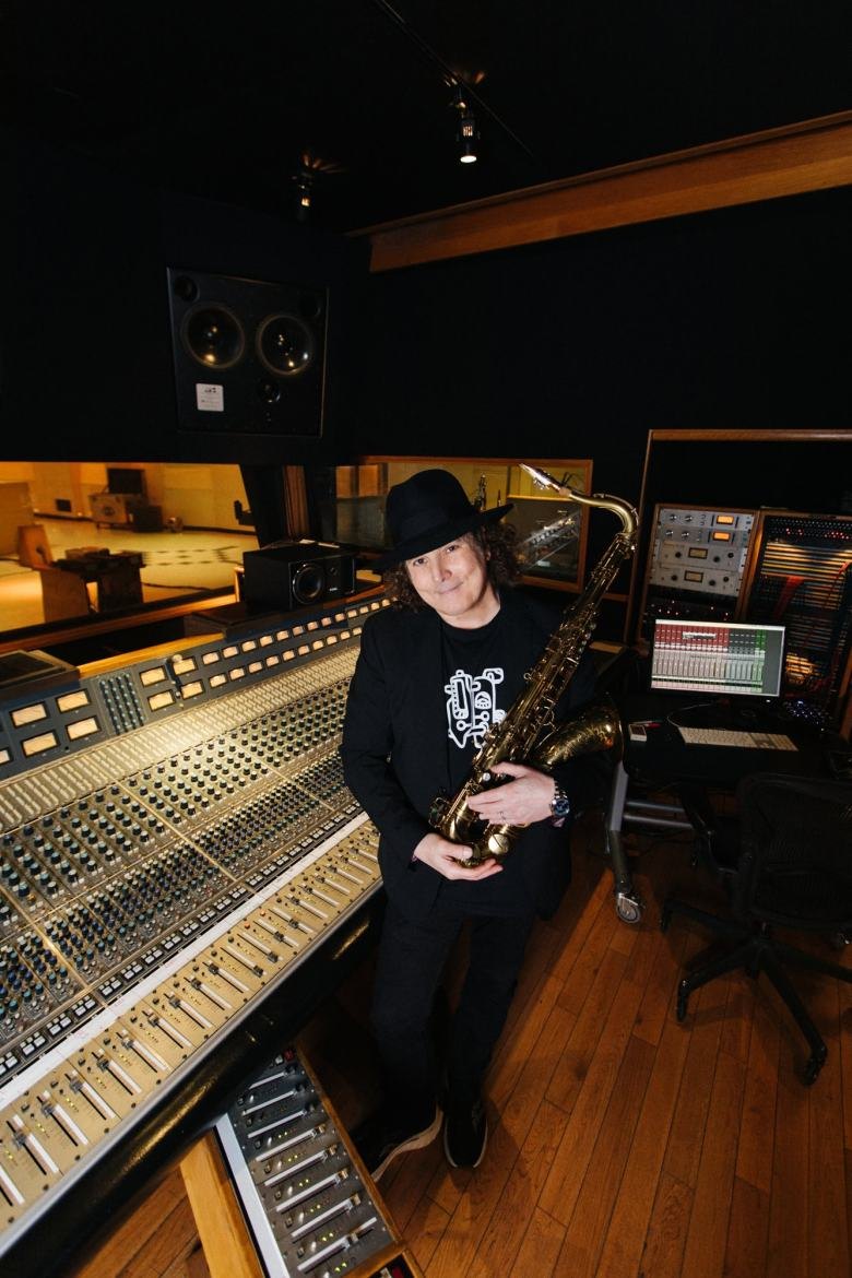 A man in a black hat holds a saxophone at a professional recording studio mixing console.