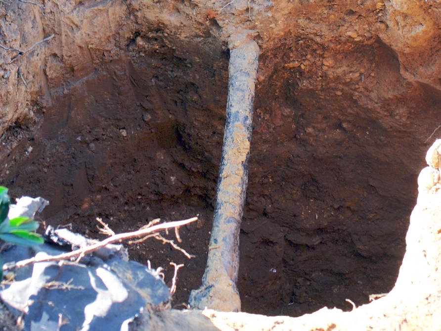 Excavated trench exposing a gray lead water pipe in dark layered soil.