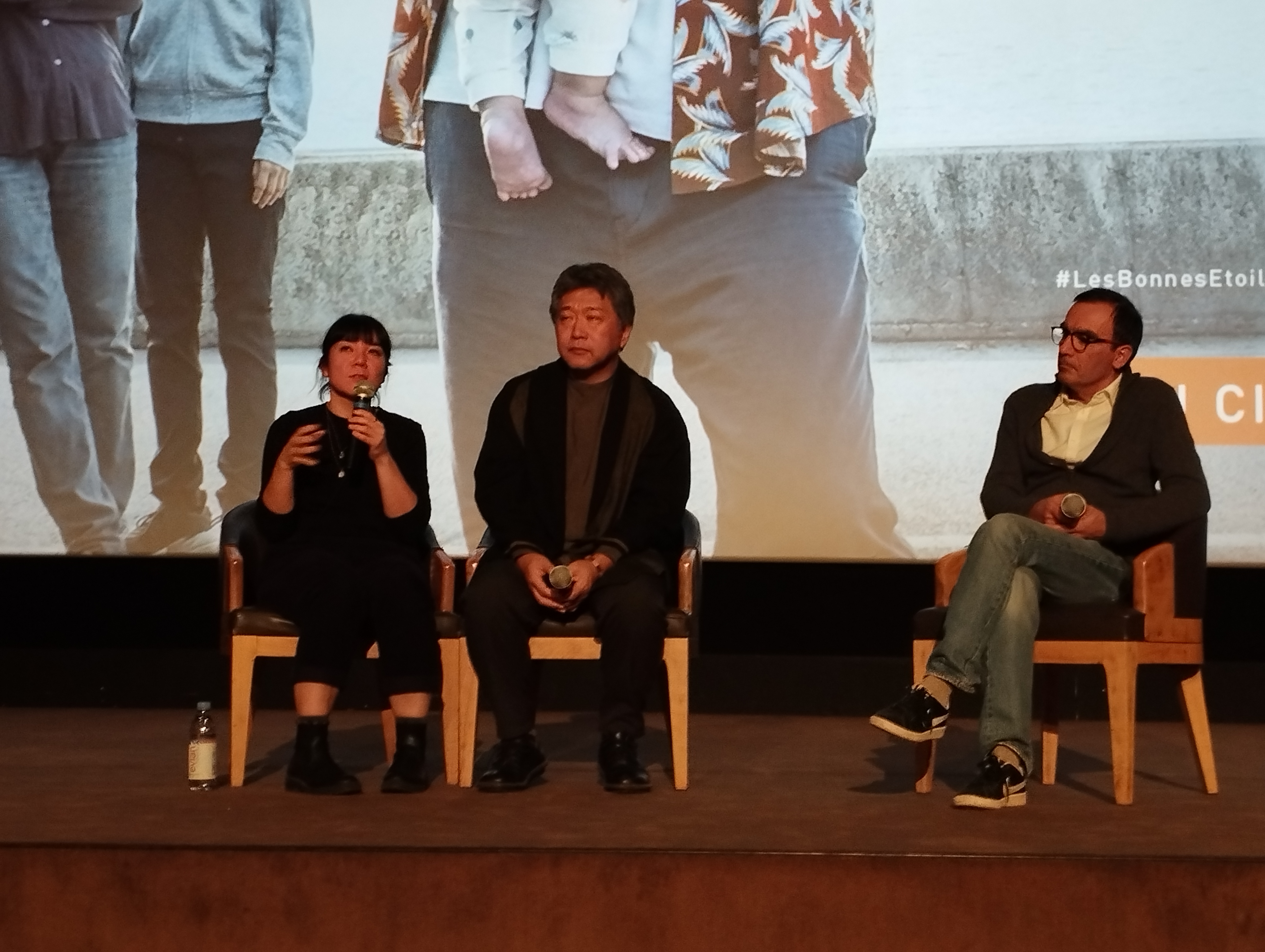 Three people seated on a stage at a film event, one speaking into a microphone with a projected screen behind them.
