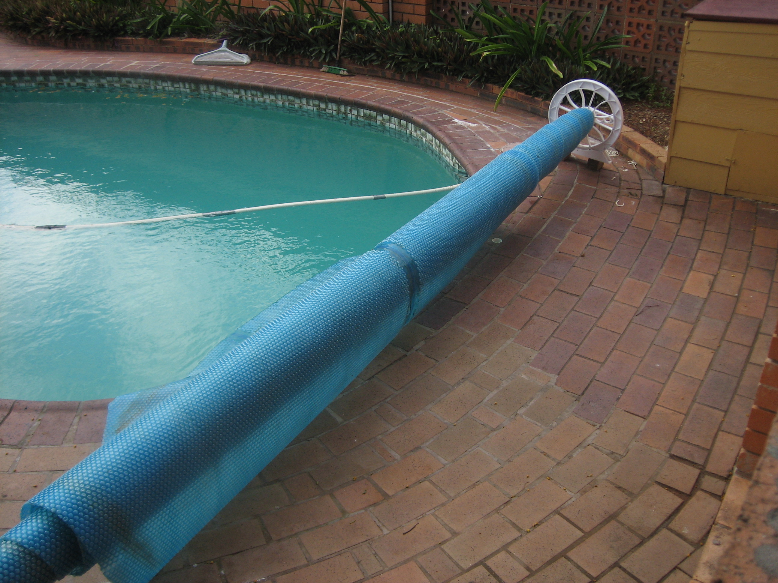 A curved residential pool with a rolled-up blue cover on a reel beside brick paving.