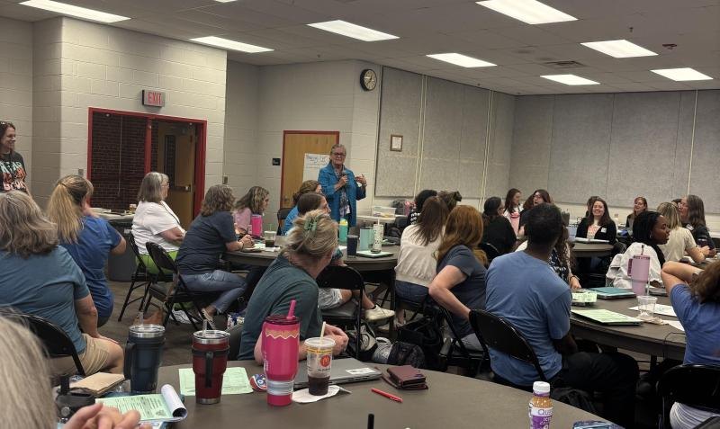 A speaker in a blue jacket addresses a large group of adults seated at round tables in a school meeting room.