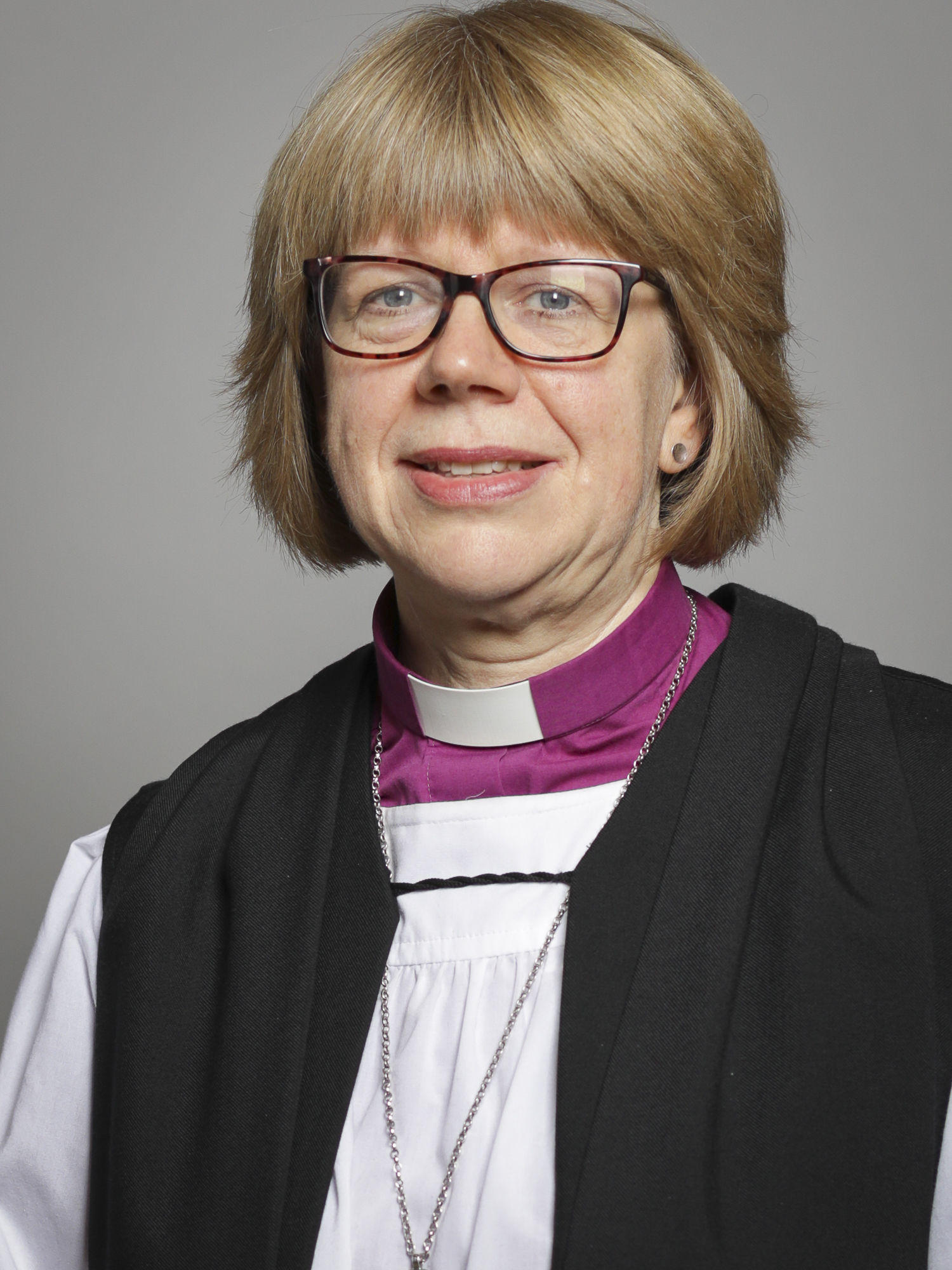 Woman in purple clerical collar, white rochet, and black chimere with red-framed glasses, posed against a grey background.