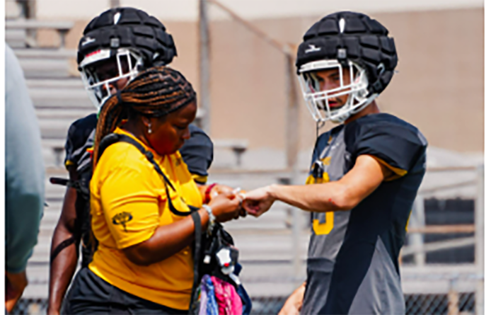 Two football players in helmets and dark jerseys talk with a staff member on a practice field sideline.