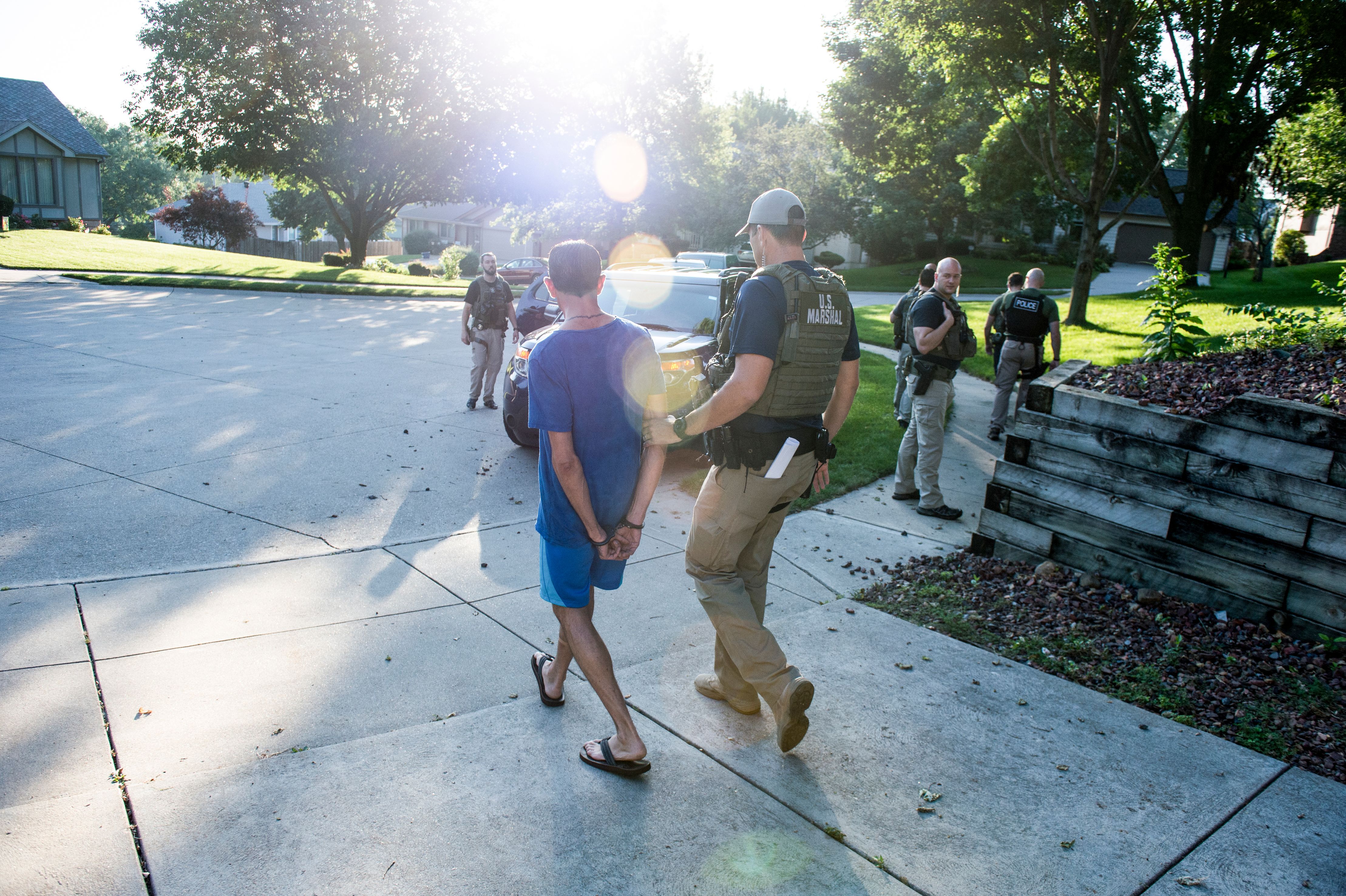 A marshal in a tactical vest escorts a handcuffed man along a sidewalk.