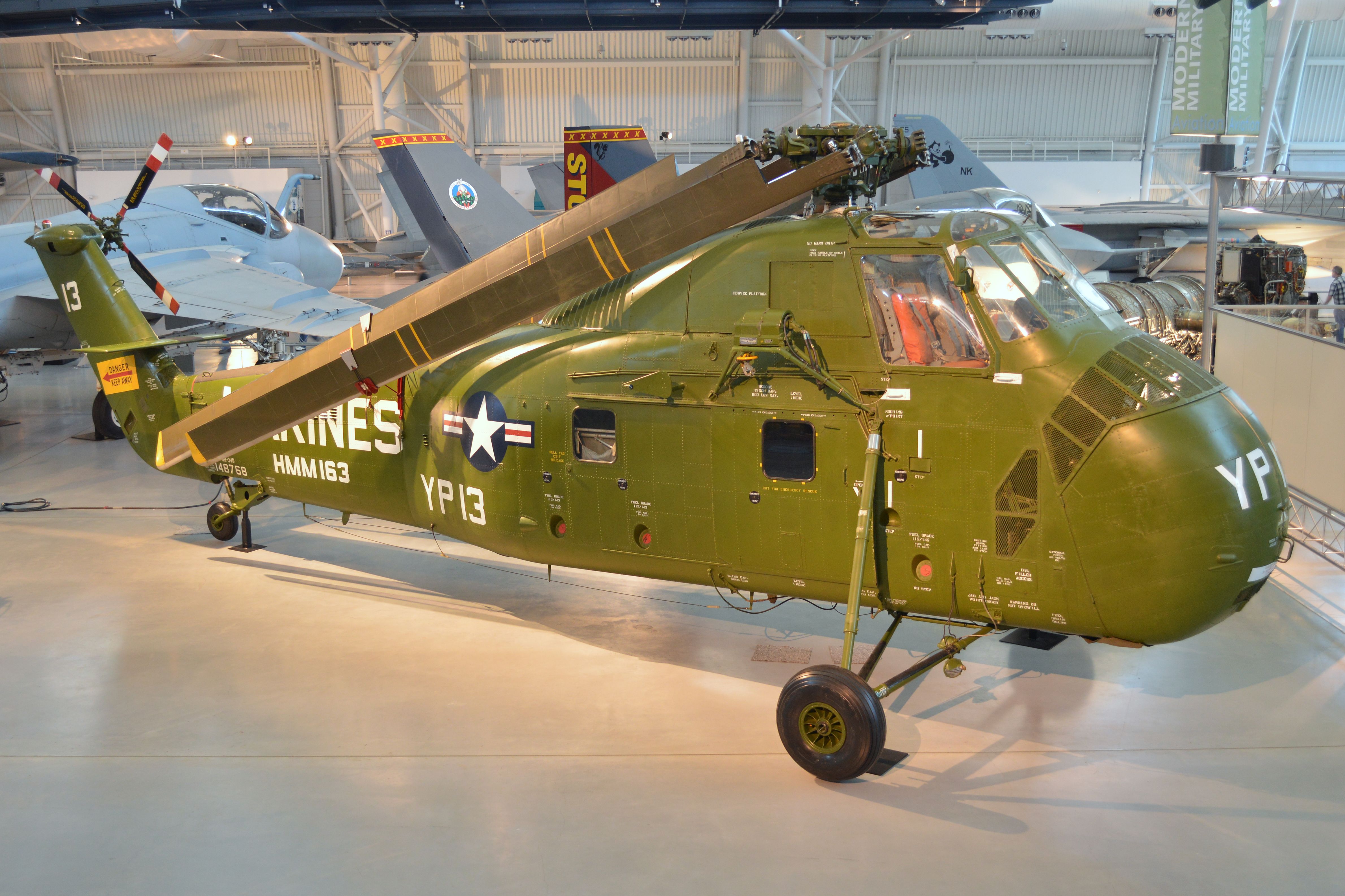 Green military helicopter on indoor display inside a museum hangar.