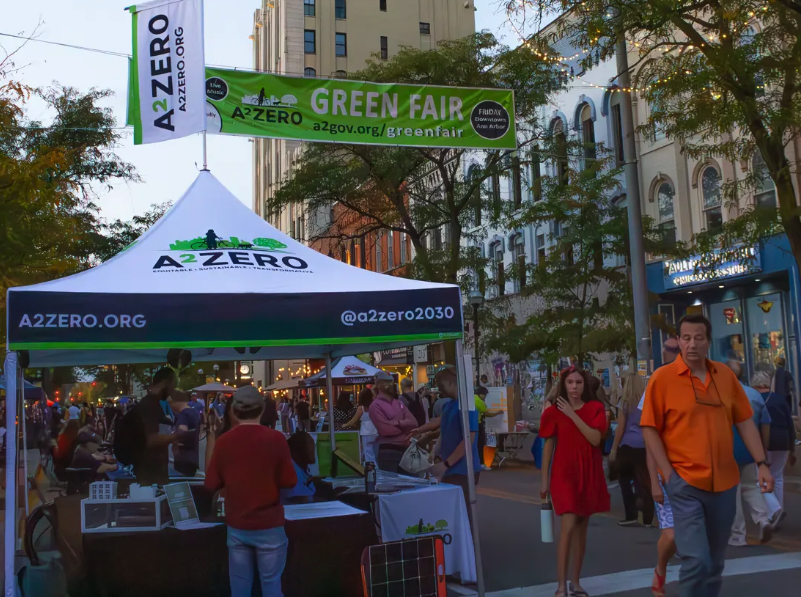 A2ZERO tent with a Green Fair banner overhead on Main Street in downtown Ann Arbor, with pedestrians walking past at dusk.