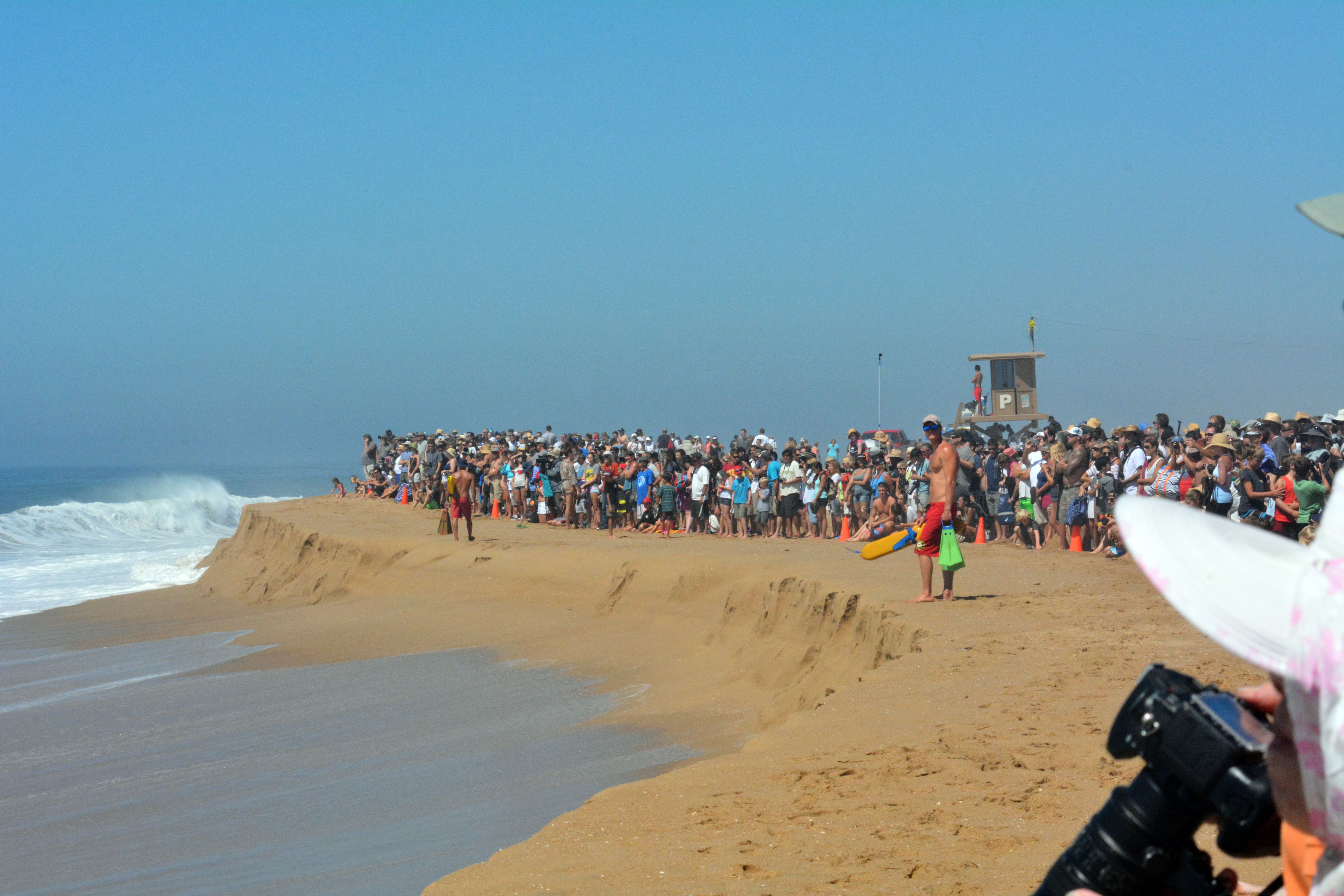 A large crowd watches powerful waves from a steep-edged beach, lifeguard tower in background.