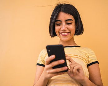 A woman smiles while looking at and using a smartphone against a yellow-orange background.