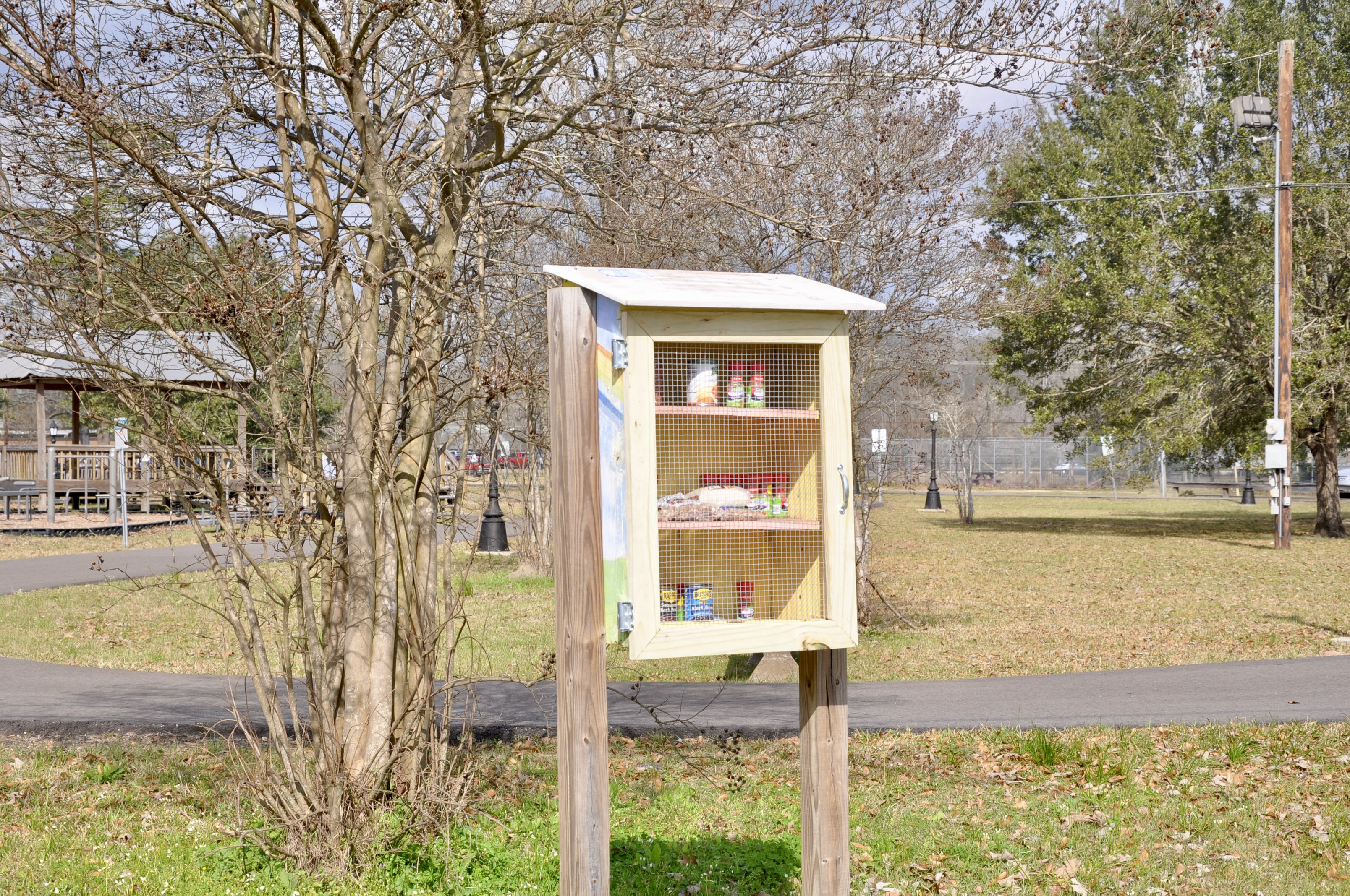 A small wooden community food pantry on posts filled with canned goods in a park setting.