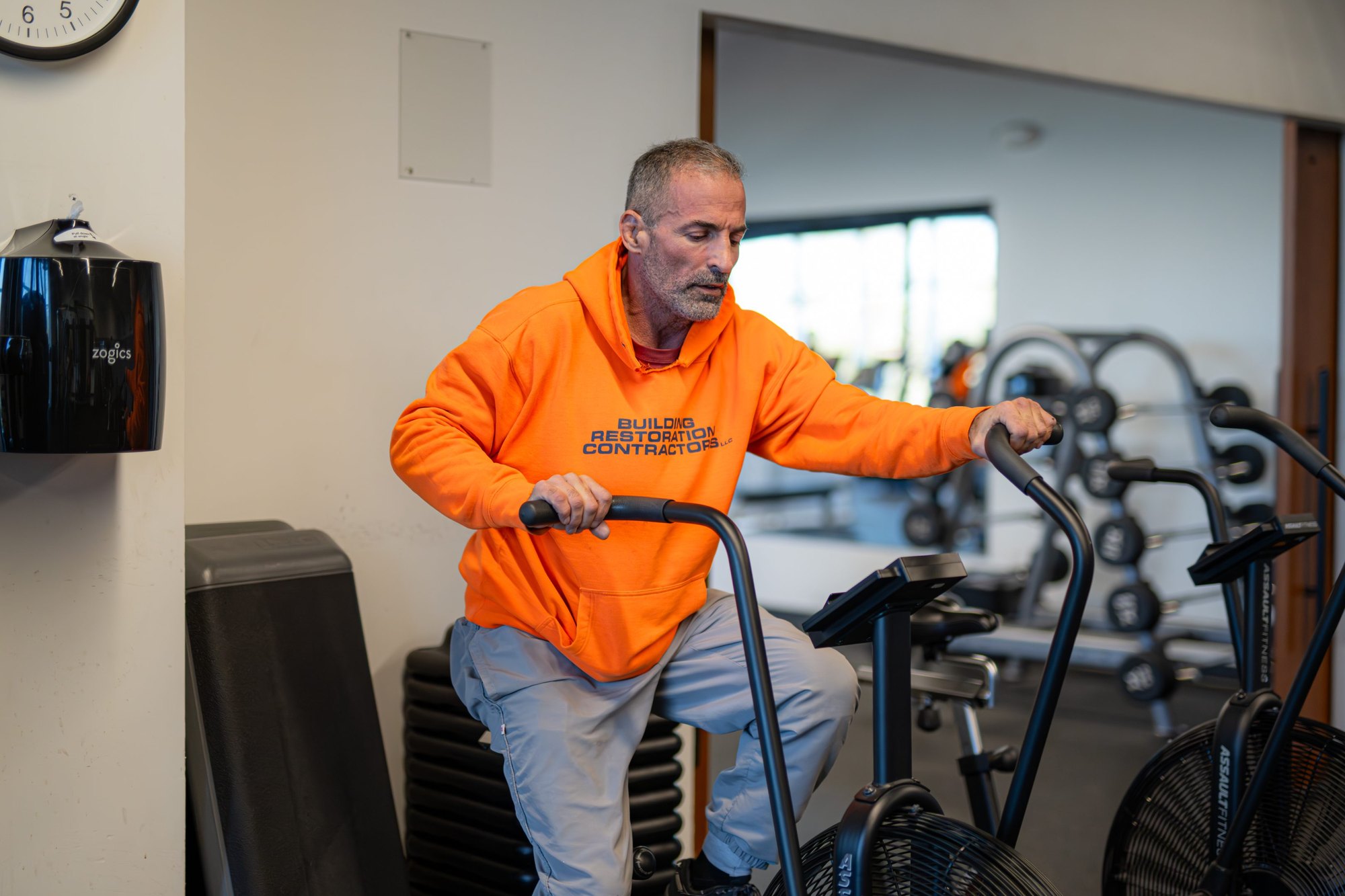 A man in an orange hoodie pedals an air bike in a gym.
